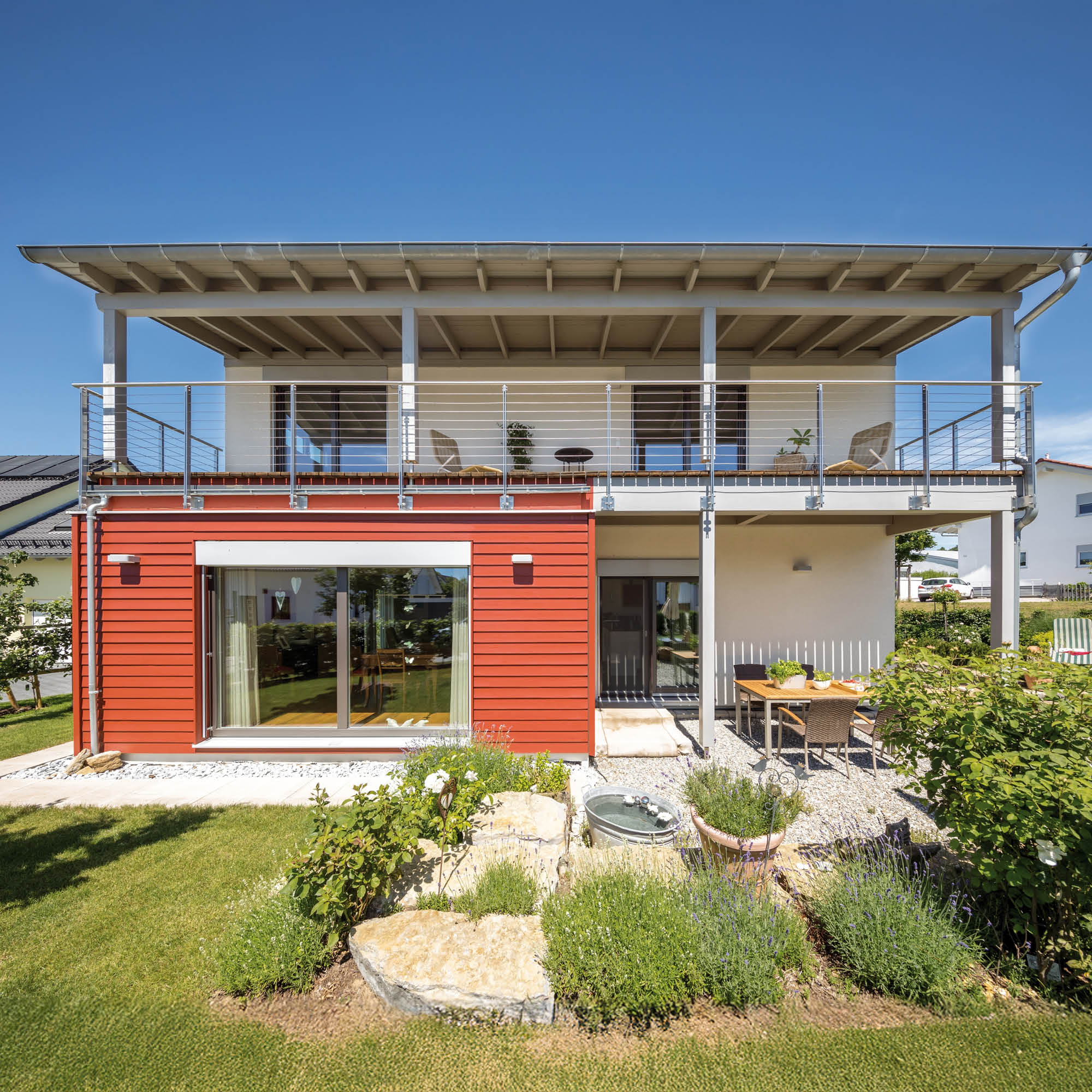 Zweistöckiges Haus mit roter Holzverkleidung im Erdgeschoss und Balkon im Obergeschoss, Garten mit Sitzbereich und Bepflanzung.