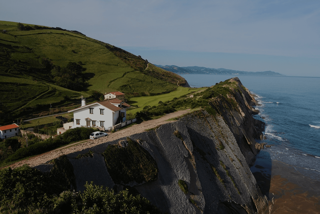A white house sits atop a dramatic cliff overlooking a tranquil ocean. Lush green hills and a coastal road are visible.
