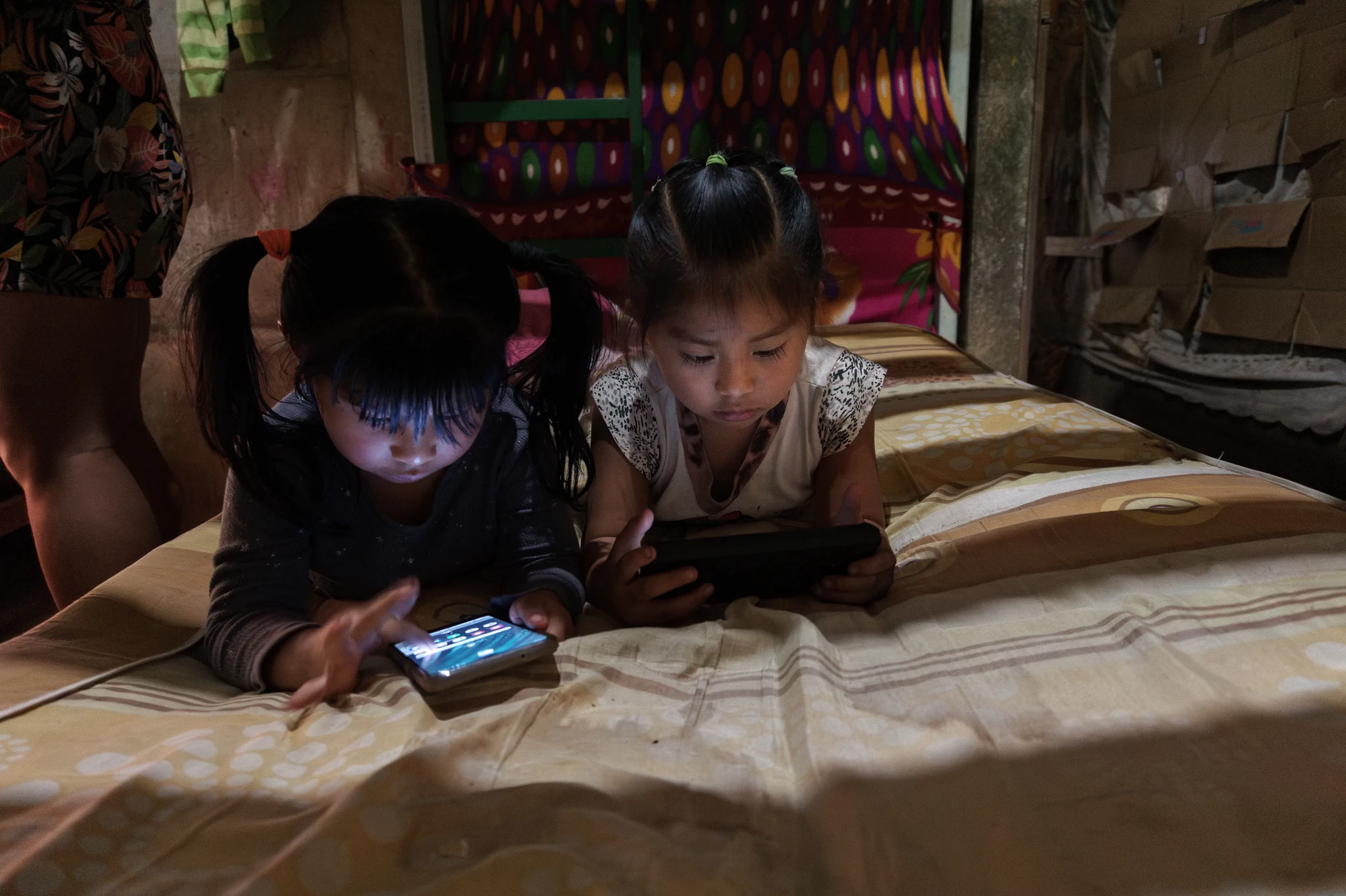 Two young girls with ponytails are lying on a bed in a dimly lit room, engrossed in playing on their smartphones. The colorful backdrop and casual setting suggest a relaxed, cozy atmosphere.
