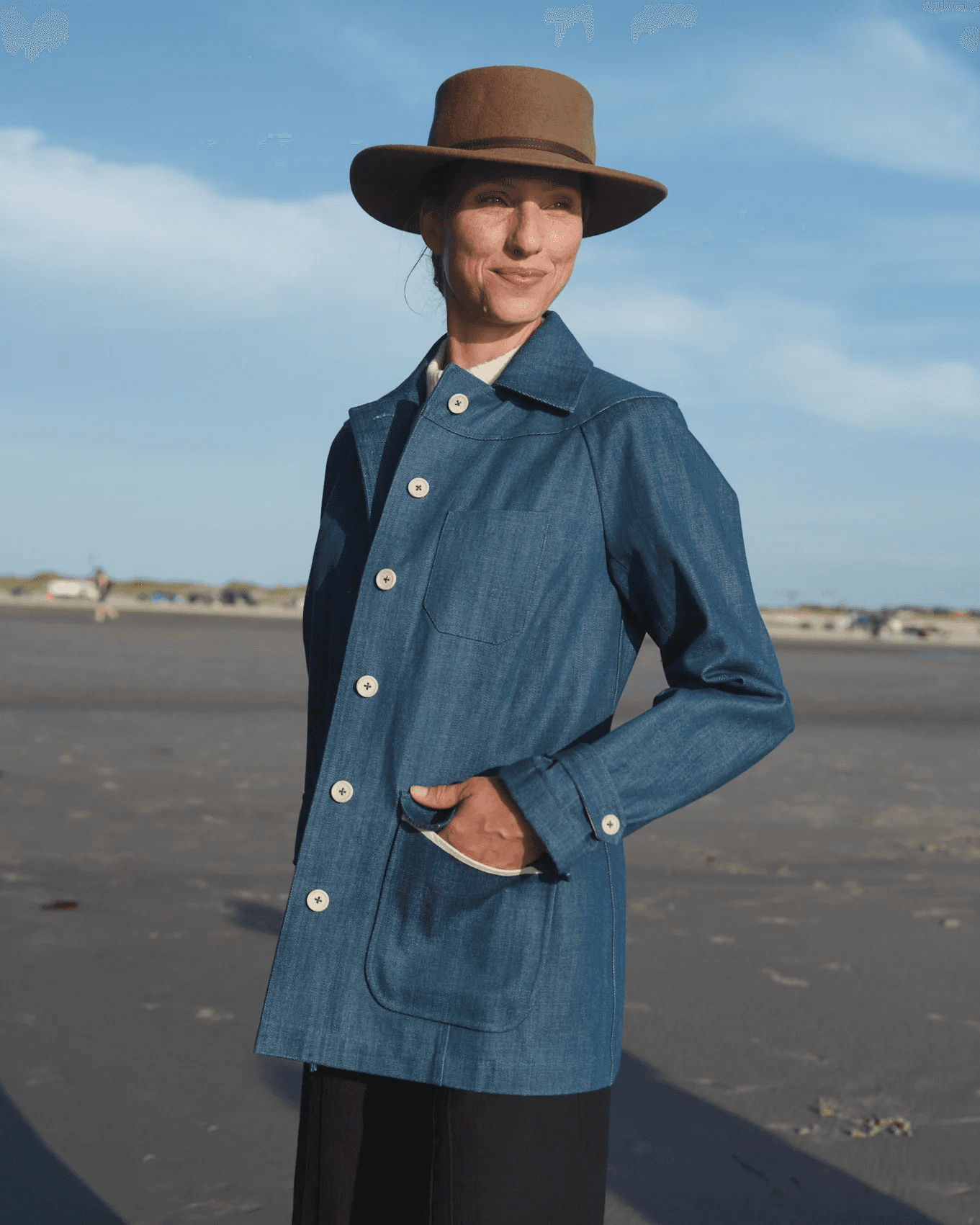 A person wearing a brown wide-brimmed hat and a blue denim jacket stands on a sandy beach under a clear blue sky, with soft sunlight casting gentle shadows.