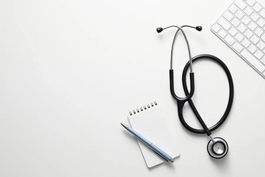 AI healthcare administration tools represented by a black stethoscope, spiral notepad, pen, and keyboard arranged neatly on a white desk surface.