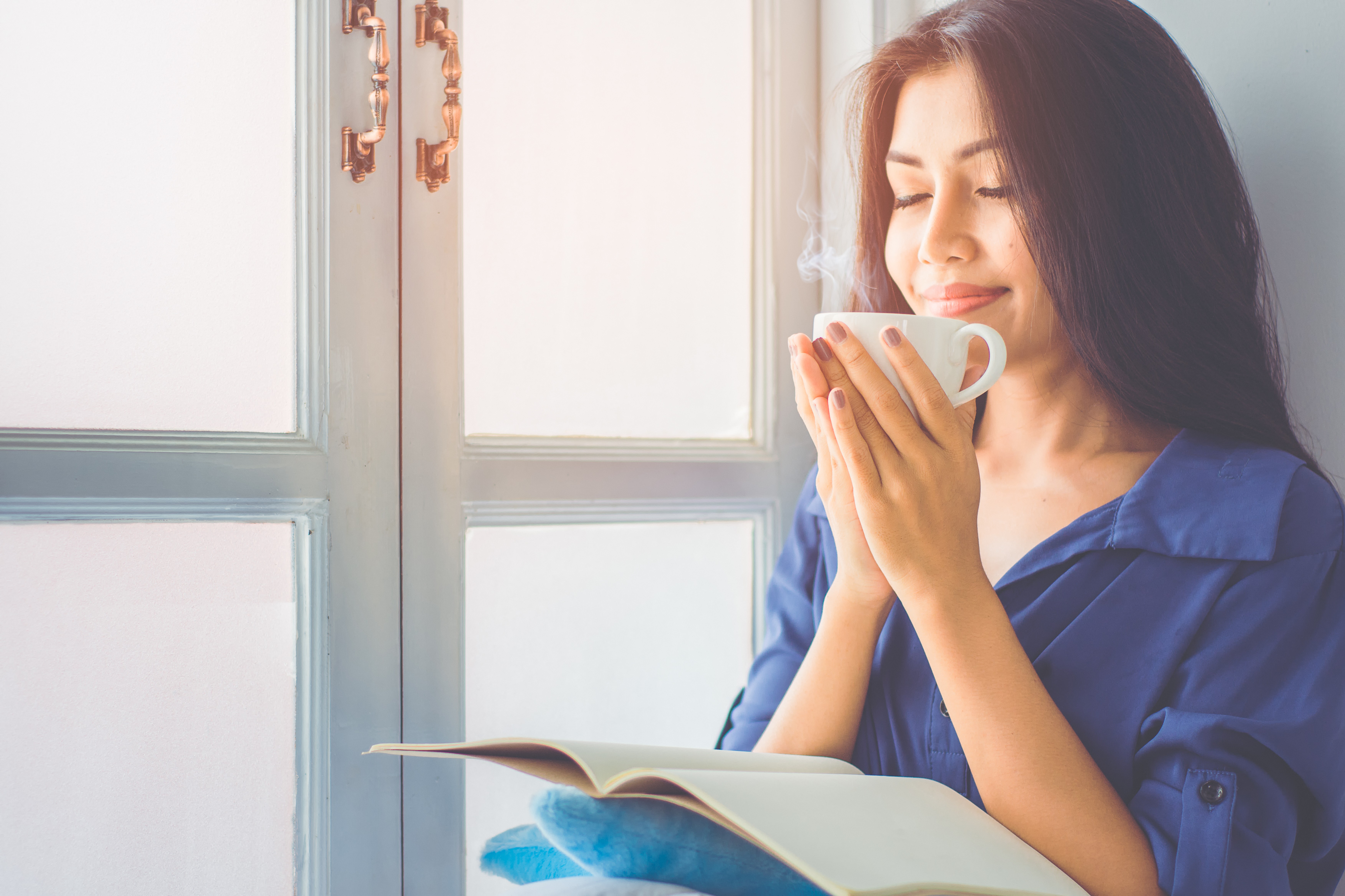 A calming pre-bed routine can help with sleep. In this image a woman in a blue shirt relaxes with a herbal tea and a book.