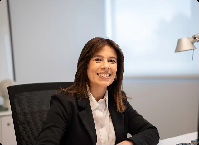 Mulher empresária sorrindo em um ambiente de escritório moderno, vestida com blazer preto e camisa branca, transmitindo confiança e profissionalismo.
