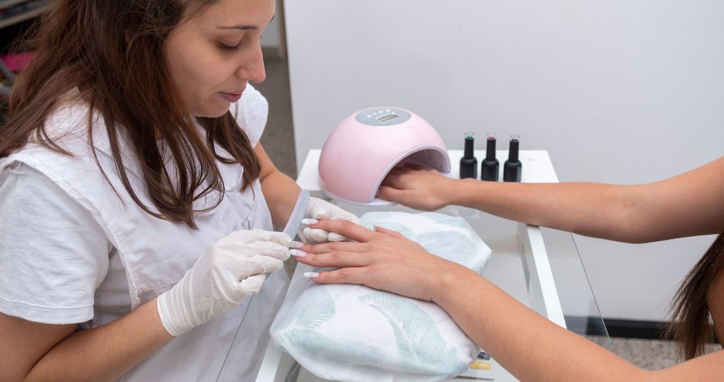 Nail technician filing a client's nails under a UV lamp at a salon.