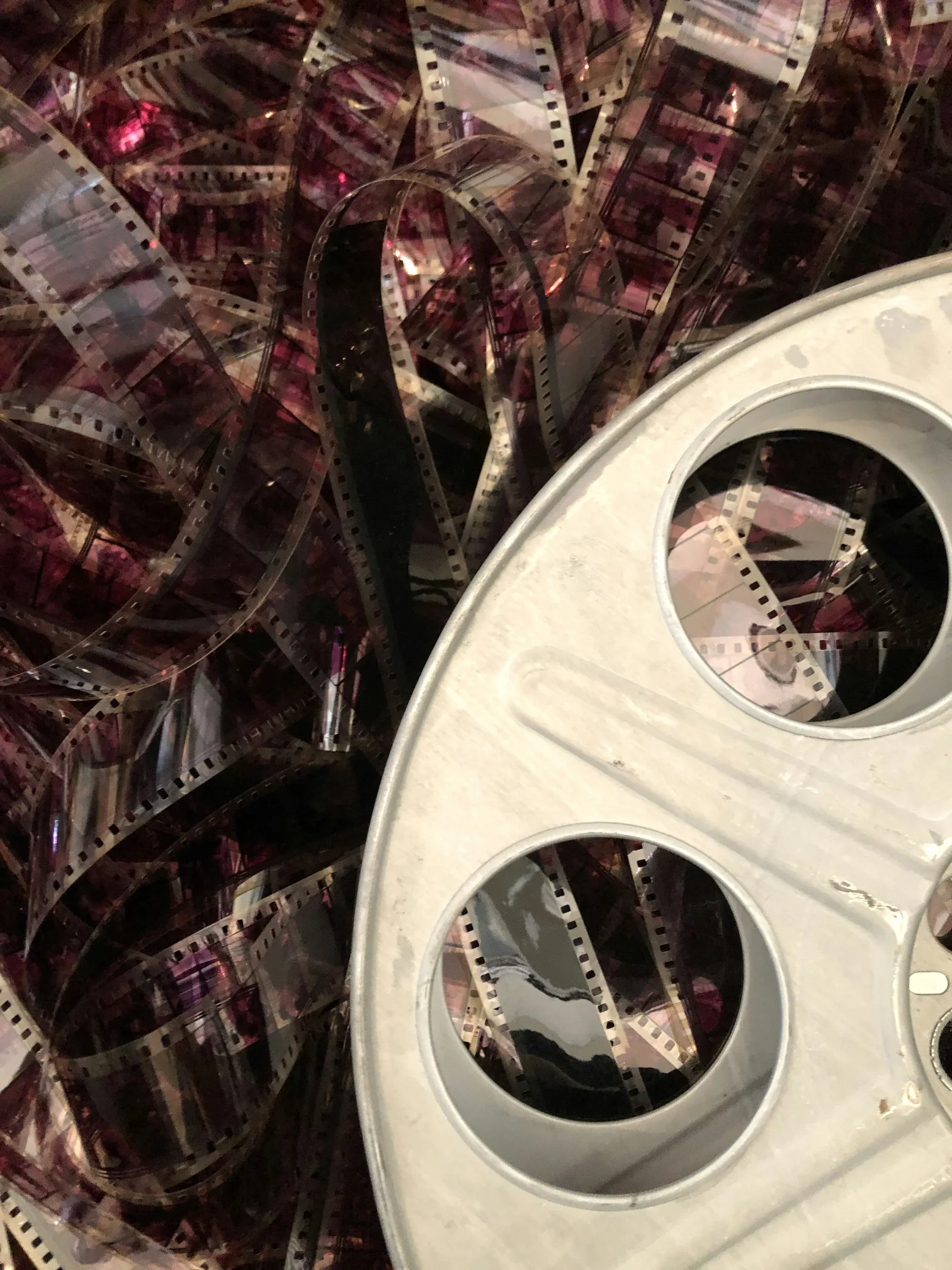A collection of tangled film strips in various shades of pink and black is displayed alongside a metallic film reel on a flat surface.