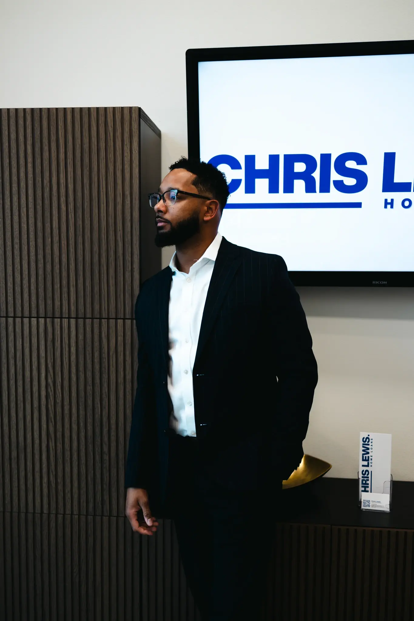 Chris Lewis stands confidently in a modern office wearing a dark suit, with the Chris Lewis Home Loans logo displayed on the screen behind him. The image represents professionalism, trust, and expert mortgage guidance.
