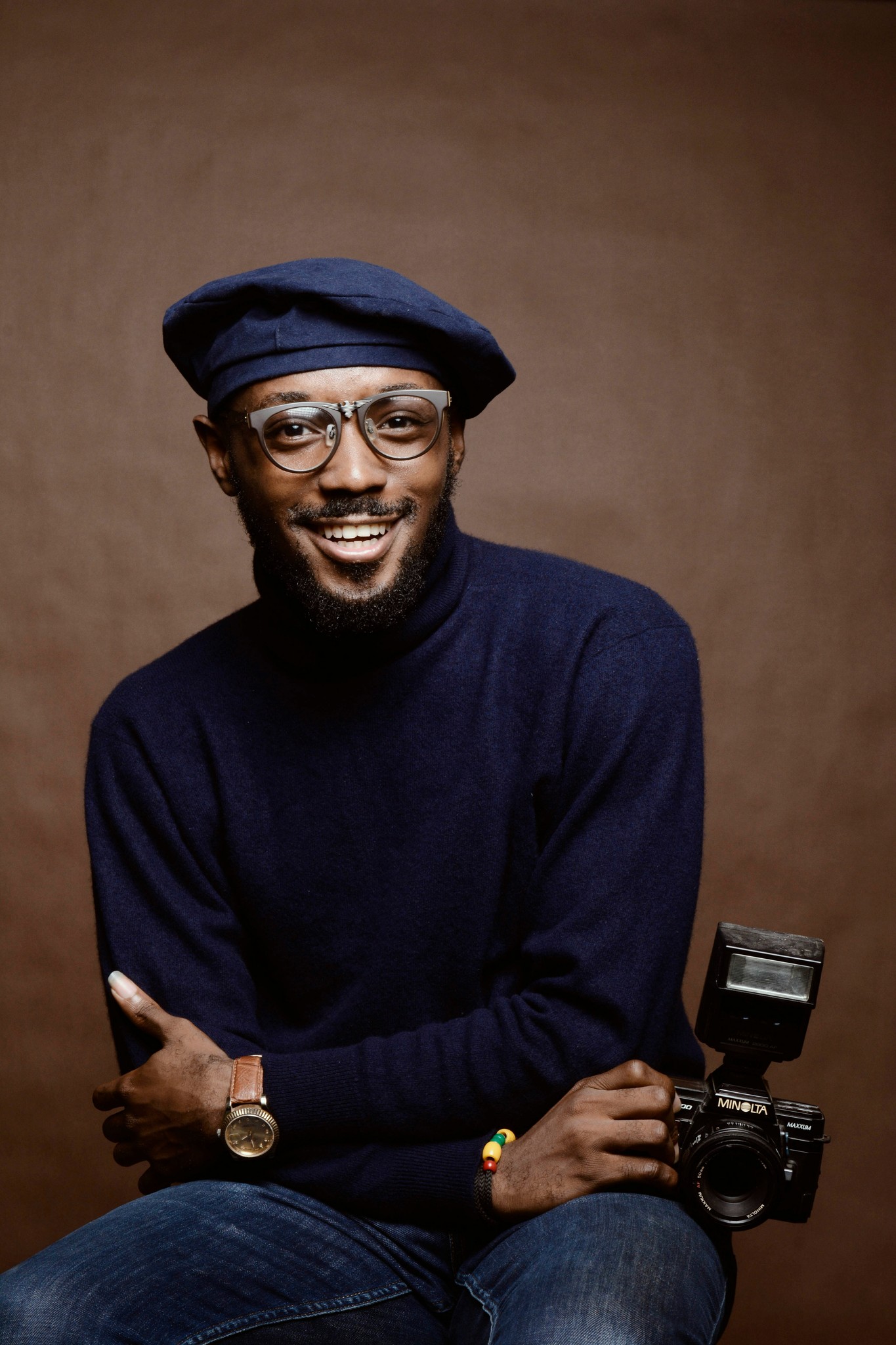 A man with glasses wearing a blue beret and jumper, smiling while holding a camera