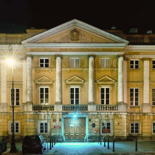 A historic building at night with large columns, lit by a street lamp. A car is parked nearby.