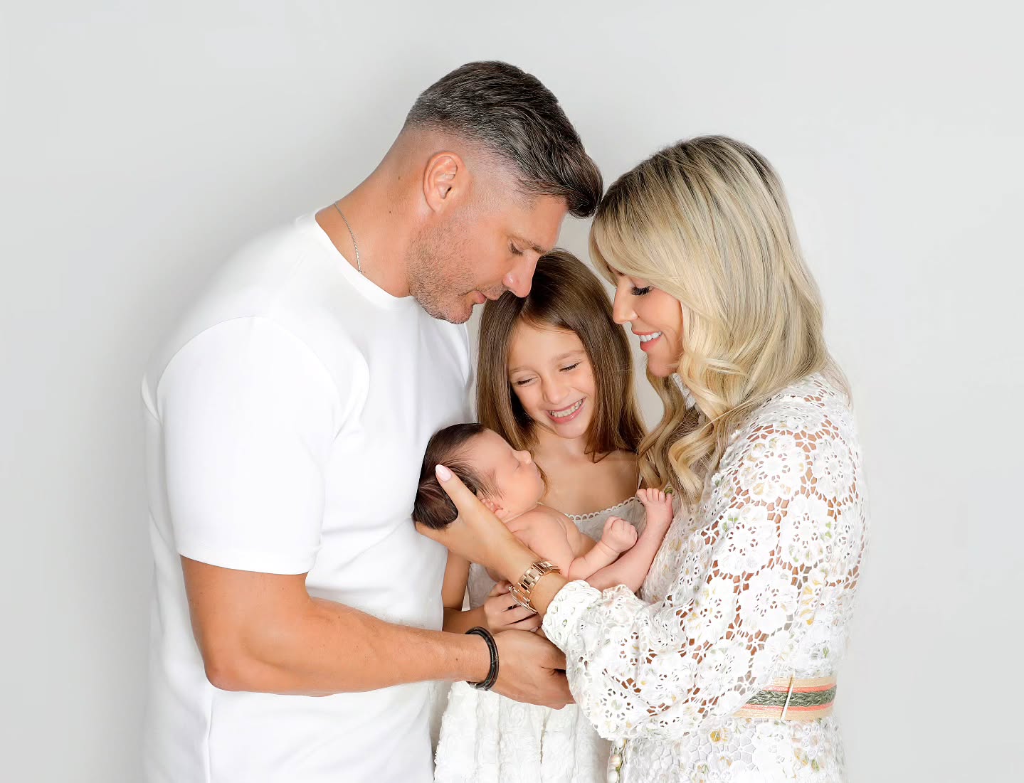 A couple pose showing the parents and an older child smiling at the baby they are holding. 