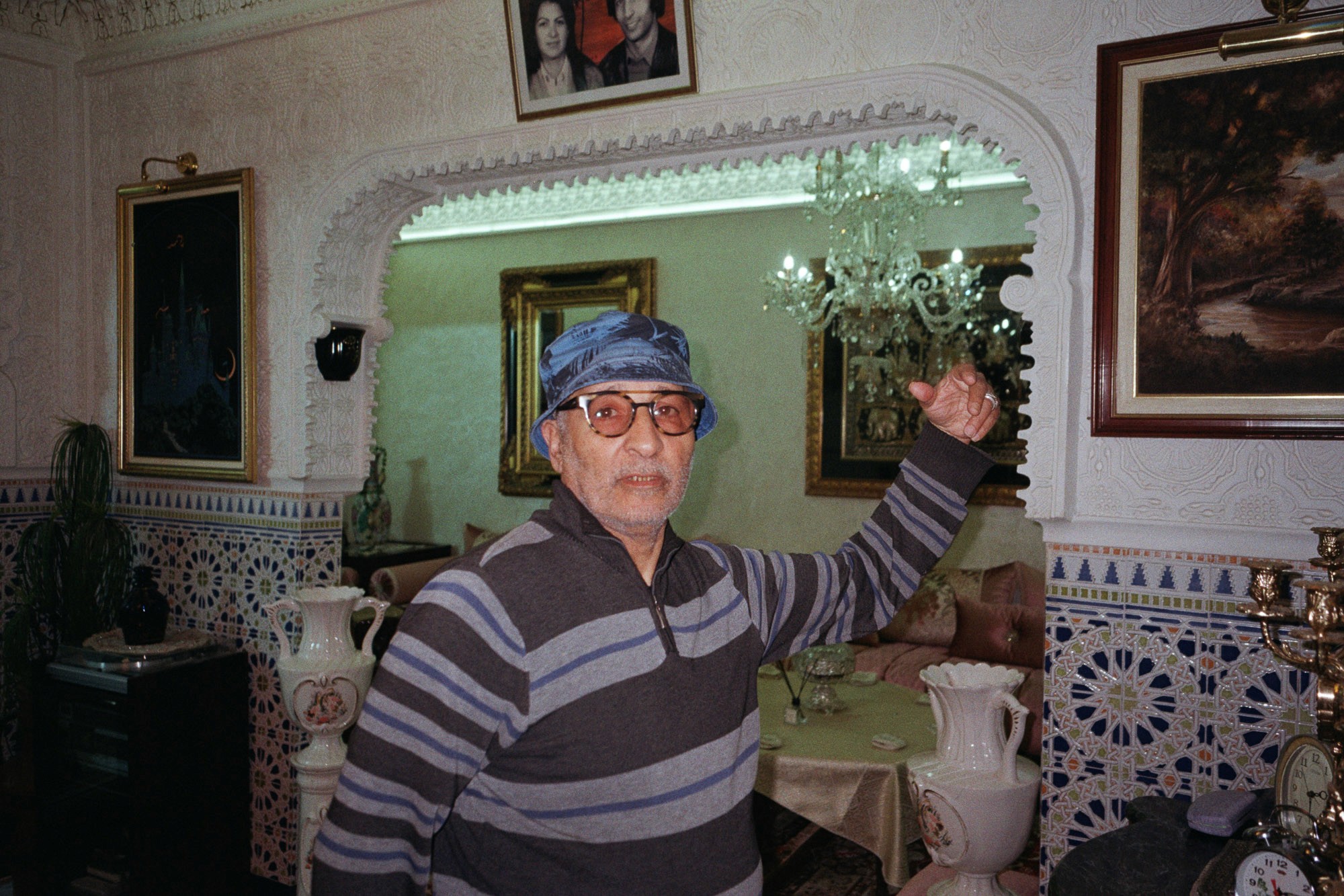 An elderly man wearing a striped sweater and blue bucket hat poses with a raised fist in an ornately decorated room featuring mosaic-tiled walls, vintage furniture, and a chandelier.