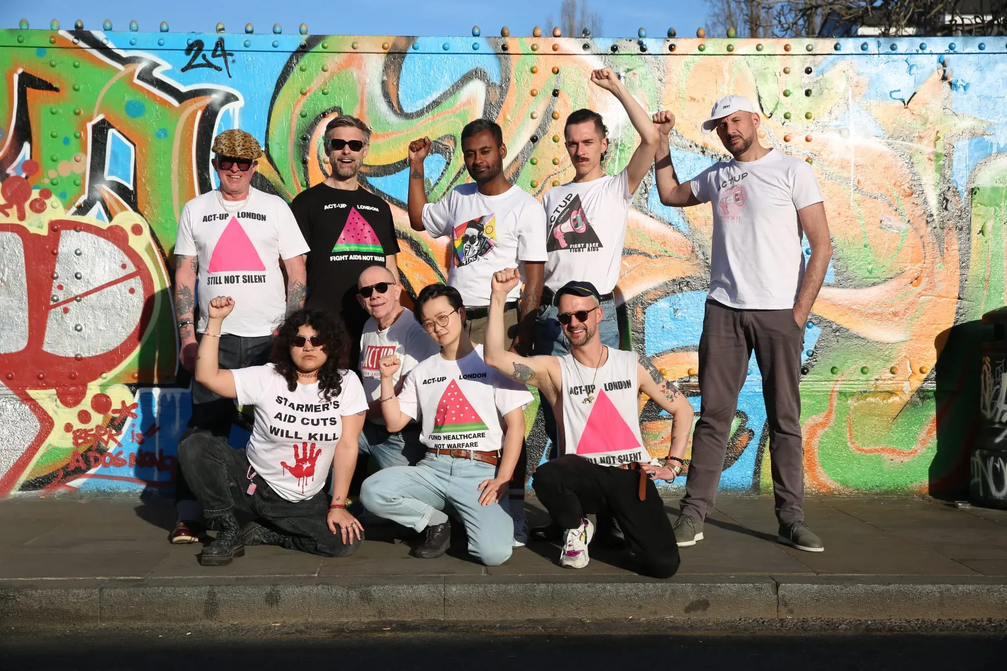 A group of present day Act Up activists wearing different Act Up t-shirts, many with pink triangles on them. The group stands or kneels in front of a brightly graffitied wall, raising a fist in a gesture of protest and defiance.
