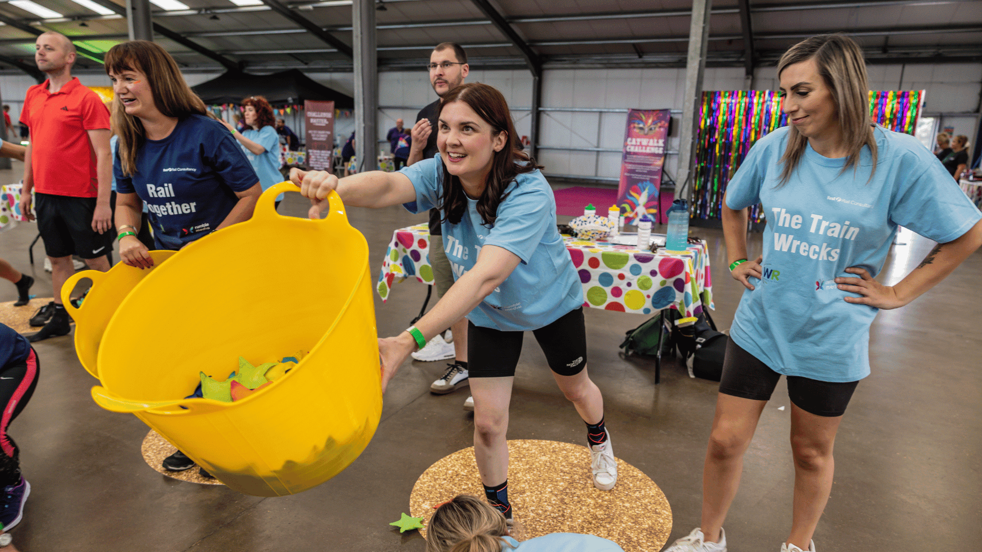 Outdoor team-building event with participants wearing matching blue shirts, taking part in inflatable obstacle courses and games, smiling and cheering, with the words 'ENTER A TEAM' in large bold white and green text across the image.