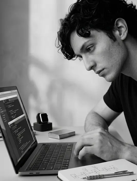 A focused individual works intently on a laptop that being the known founder Philip Heyes, surrounded by a notebook and smart devices, embodying the spirit of a traditional startup founder in a venture studio environment.