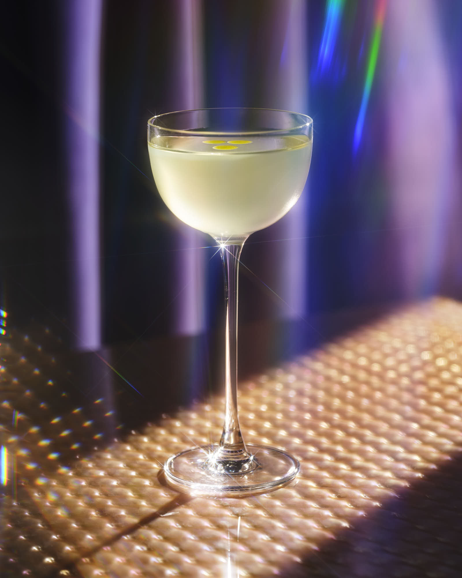 A cocktail glass filled with a light-colored drink, sitting on a lit surface with colorful lighting in the background.