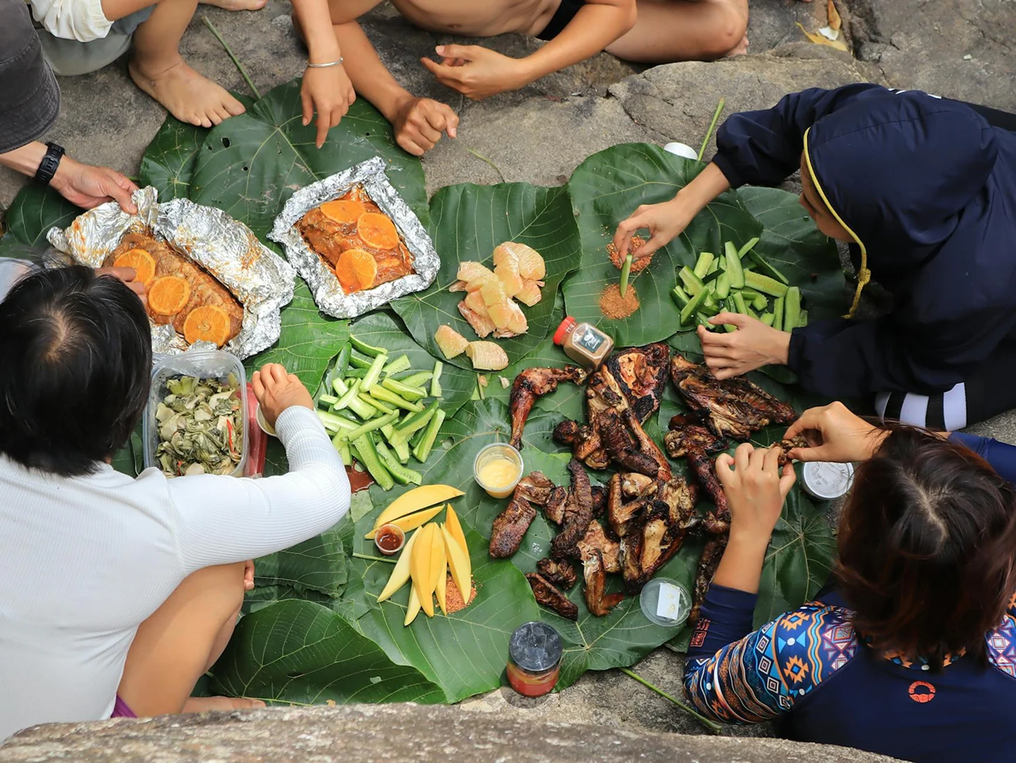 Casual beach picnic with grilled food, fruit, and snacks served on large tropical leaves.
