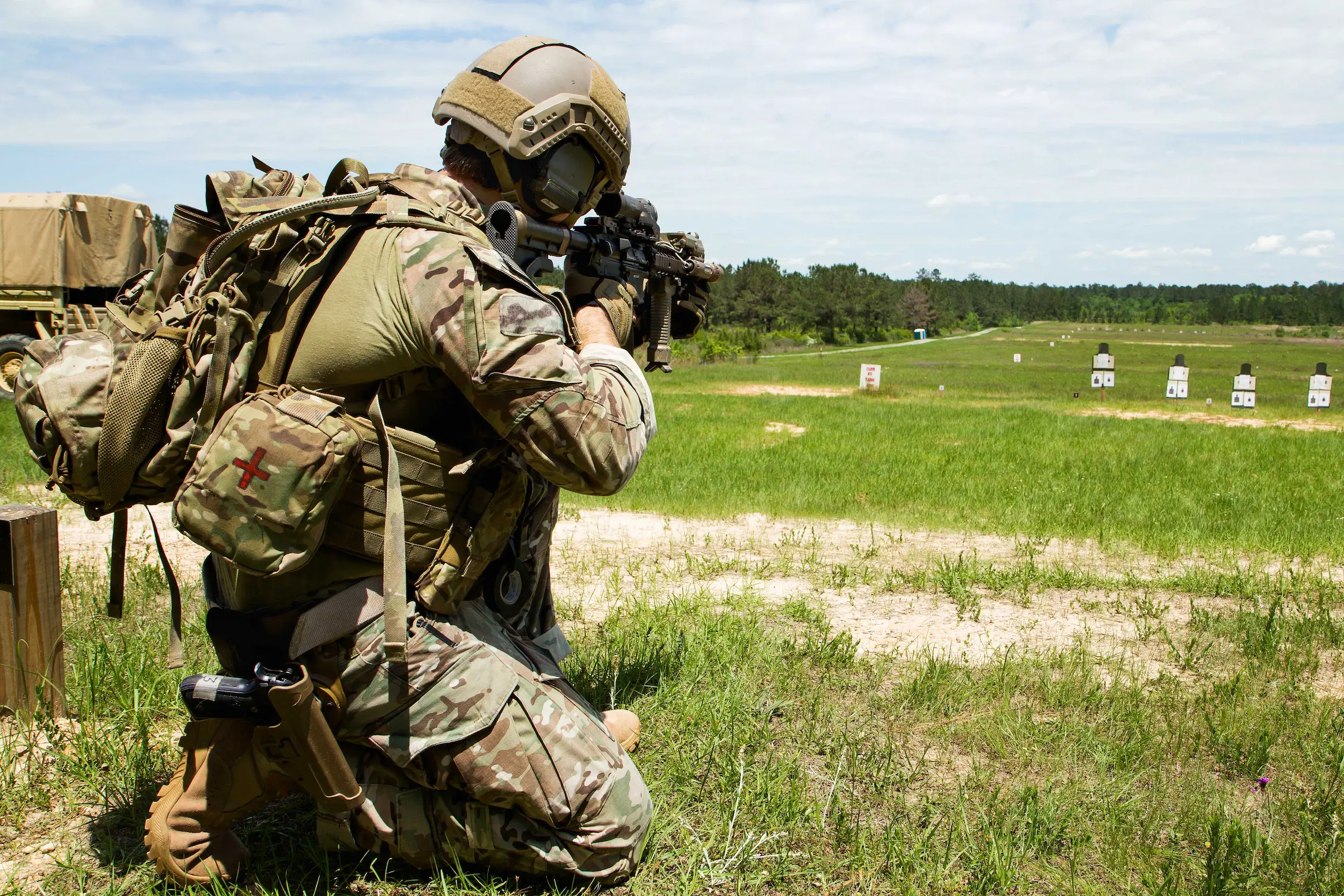 Soldier in full tactical gear kneeling on a grassy field while aiming a rifle at distant target silhouettes during a live-fire training exercise.