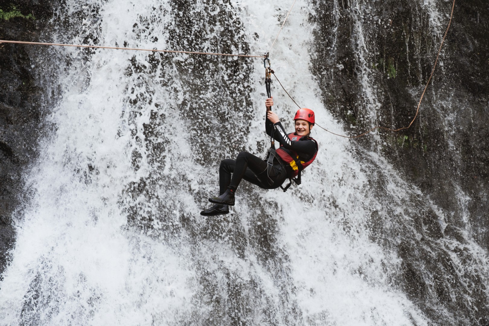 Participant wearing a red helmet suspended on a zip line in front of a cascading waterfall during a gorge adventure in North Wales.