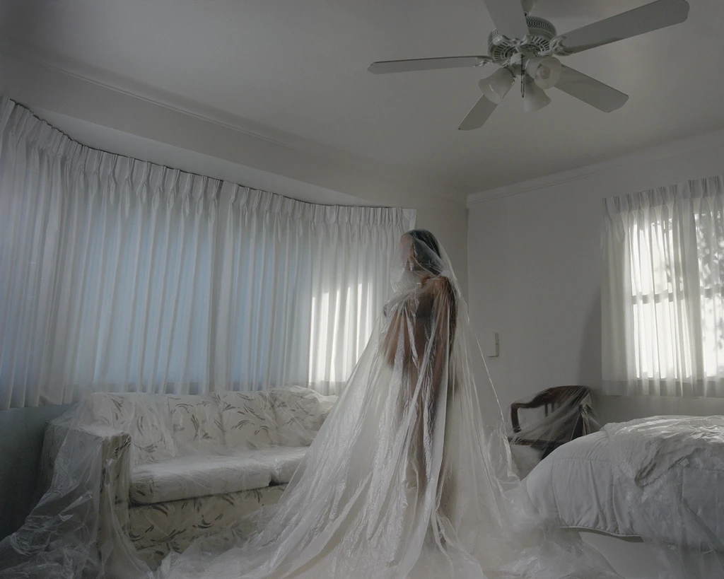 A pregnant woman stands amongst furniture draped in translucent plastic sheeting, image by Gearld Canaday IV.