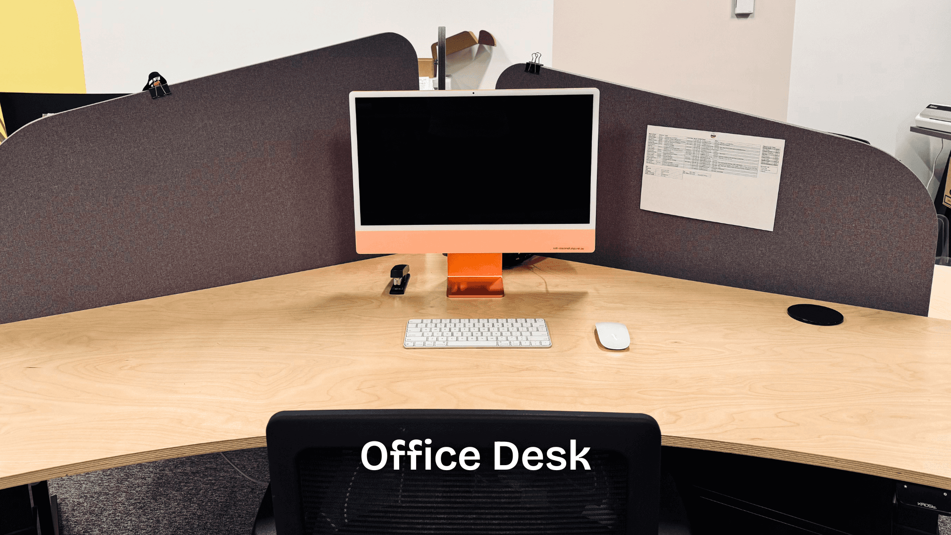 A tidy corner office workstation featuring an orange iMac, keyboard, mouse, and a stapler, set on a light timber desk with privacy screens on three sides.