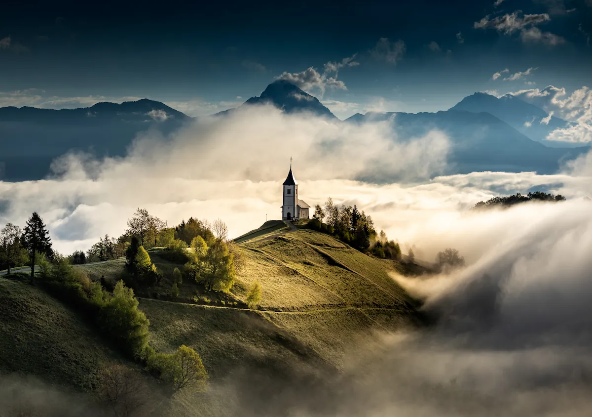 A breathtaking view of the Church of St. Primus and Felician perched on a green hilltop in Jamnik, Slovenia, emerging from a thick sea of morning fog. The scene is set against the dramatic backdrop of the Kamnik-Savinja Alps, with the sun's low light catching the rolling mist and highlighting the church's silhouette under a moody, cloud-streaked sky.