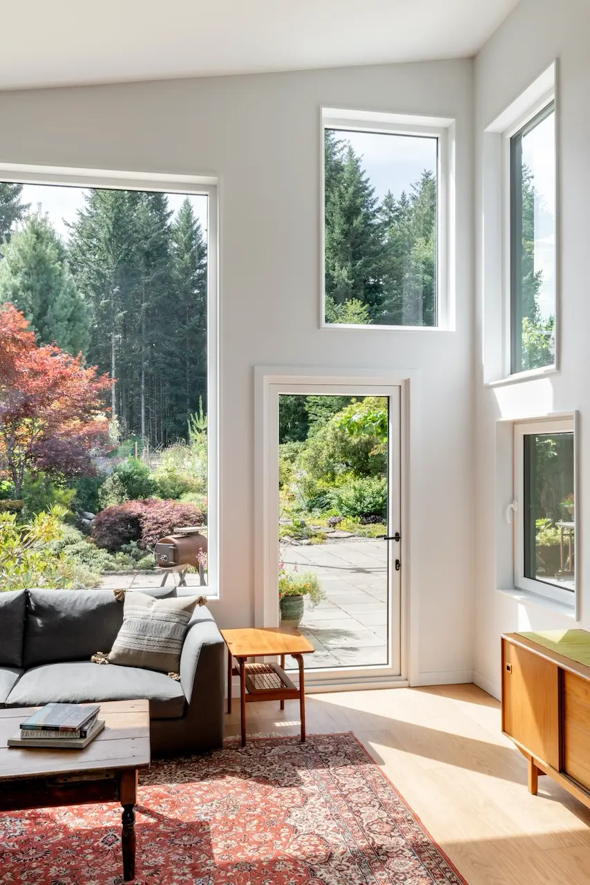 Modern two-story home with large glass windows, warmly lit interiors, and a balcony. Surrounded by trees and a manicured lawn at dusk. Cozy ambiance.