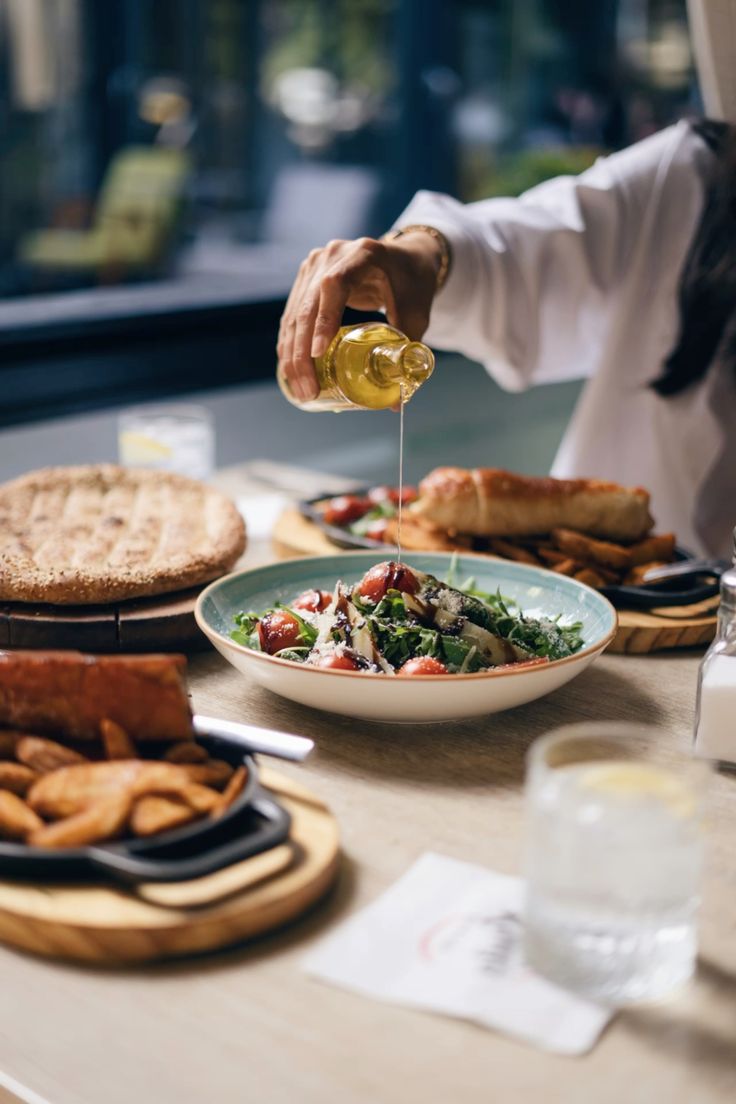 Chef finishing a fresh salad with a precise pour of dressing, representing the patience, balance, and technique centric approach that defines Dhoom’s contemporary Indian cooking.