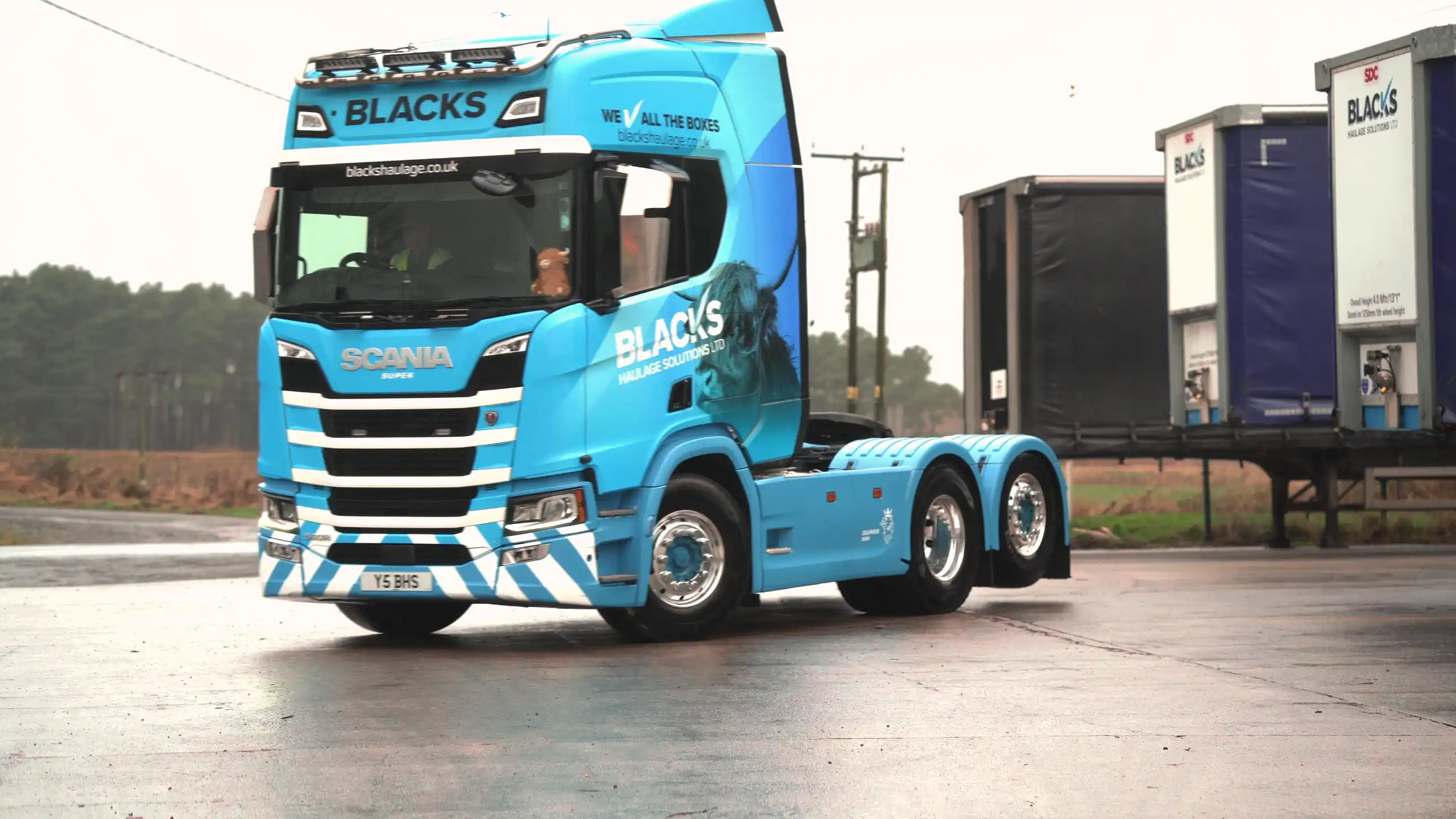 A bright blue Scania truck with "BLACKS" branding drives through an industrial area. Nearby, there are large black and white shipping containers.