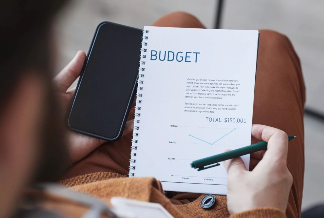 A person holding a phone and looking at a physical copy of a budget. 