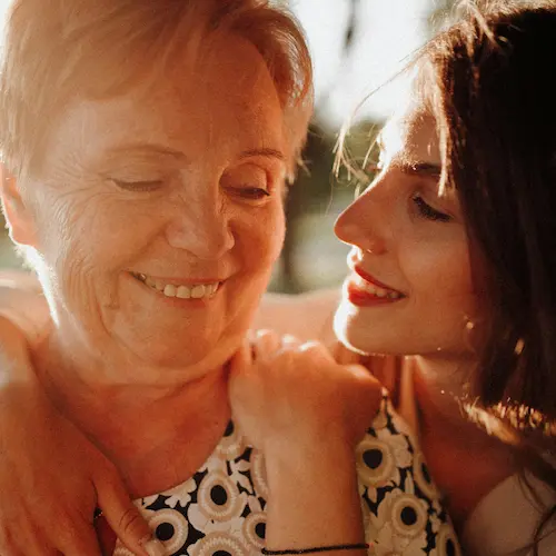 Older woman and younger woman sharing a warm, close moment together, both smiling in soft natural light.