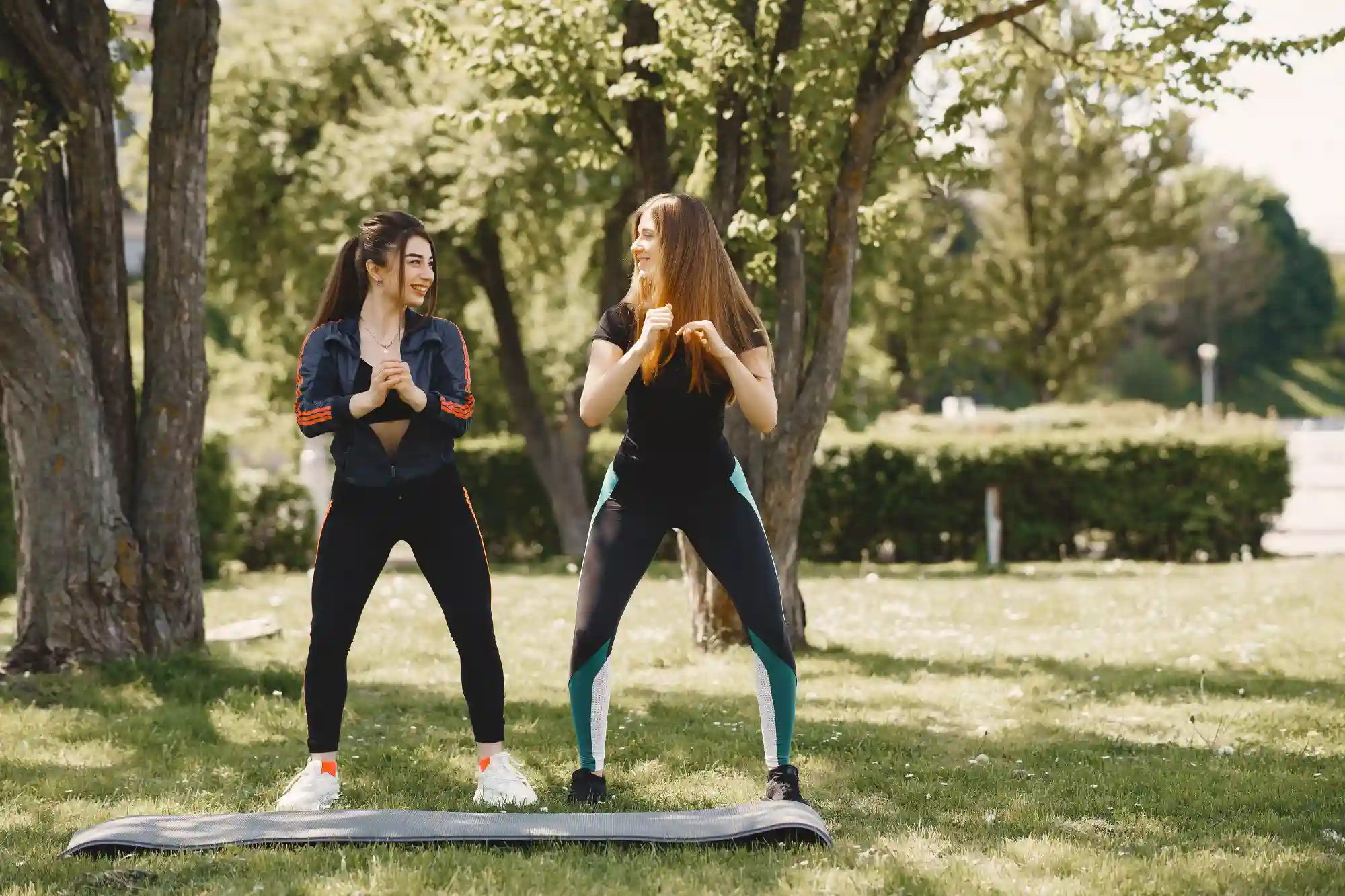 Two young women in athletic wear smiling and performing squats together on a yoga mat in a sunny park.
