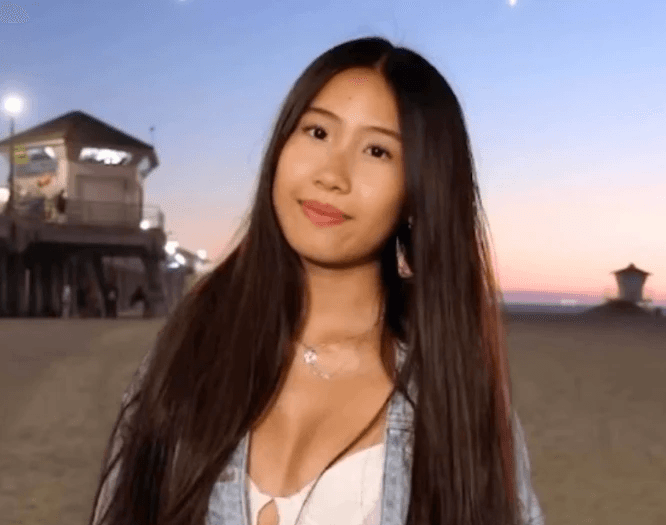 A person with curly hair and glasses smiles at the camera, seated in a cozy workspace with warm lighting.