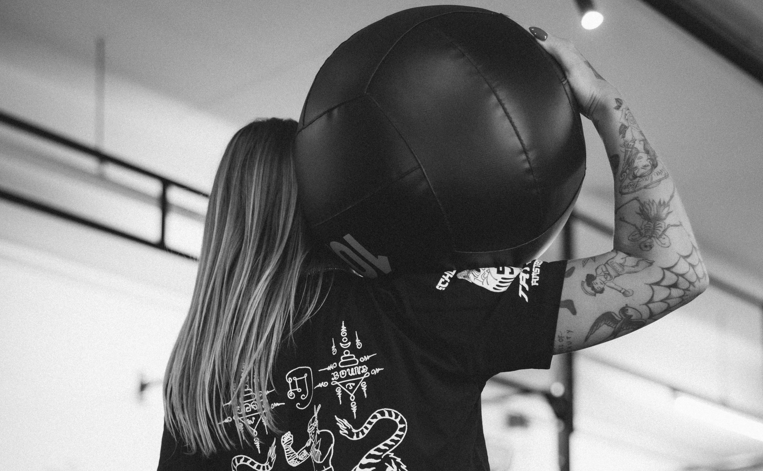 A person with long hair holds a large black ball overhead in a gym setting. The image is in black and white.