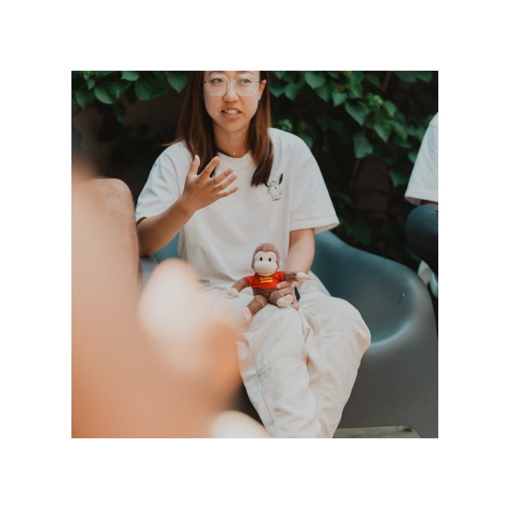 A woman sits on a chair outdoors, gesturing while speaking. She holds a toy monkey wearing a red shirt. Green leaves form the background, conveying a casual, relaxed setting.