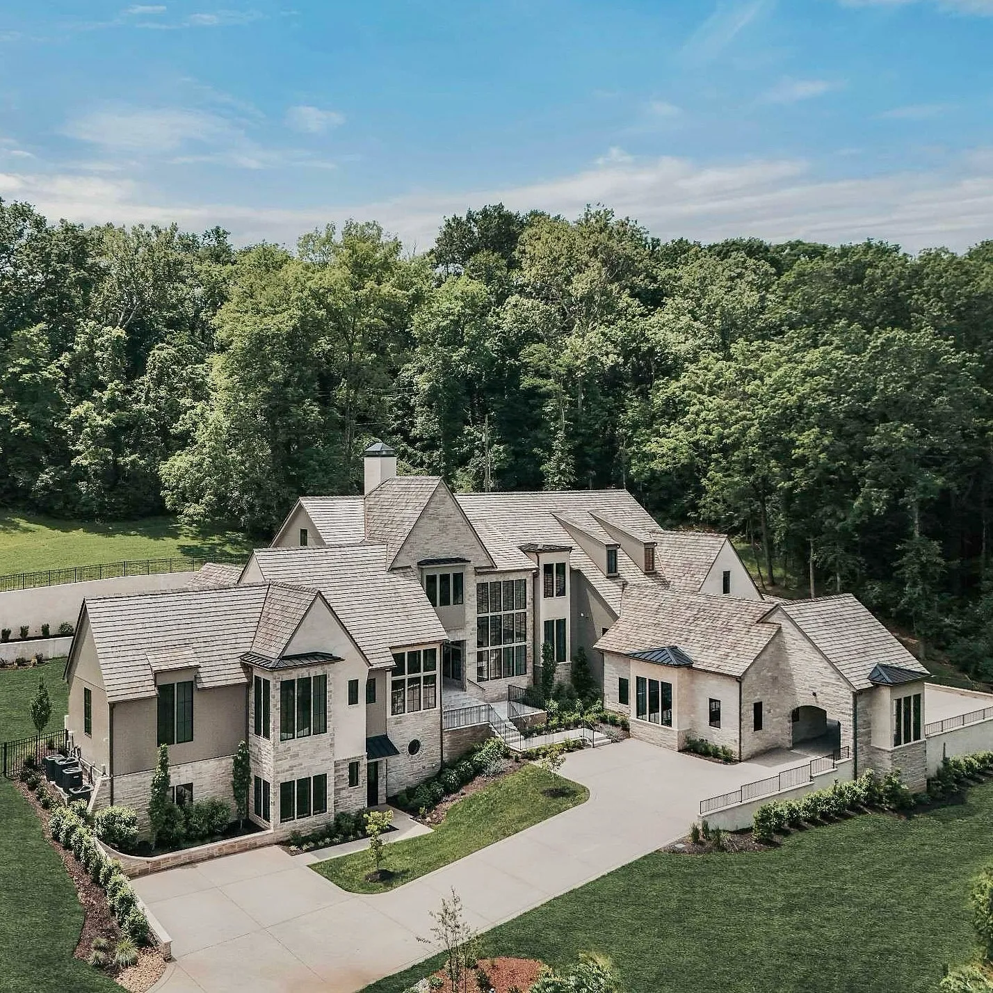 An aerial view of a large, luxurious house with a shingled roof, surrounded by lush green trees and a well-maintained lawn with a driveway.