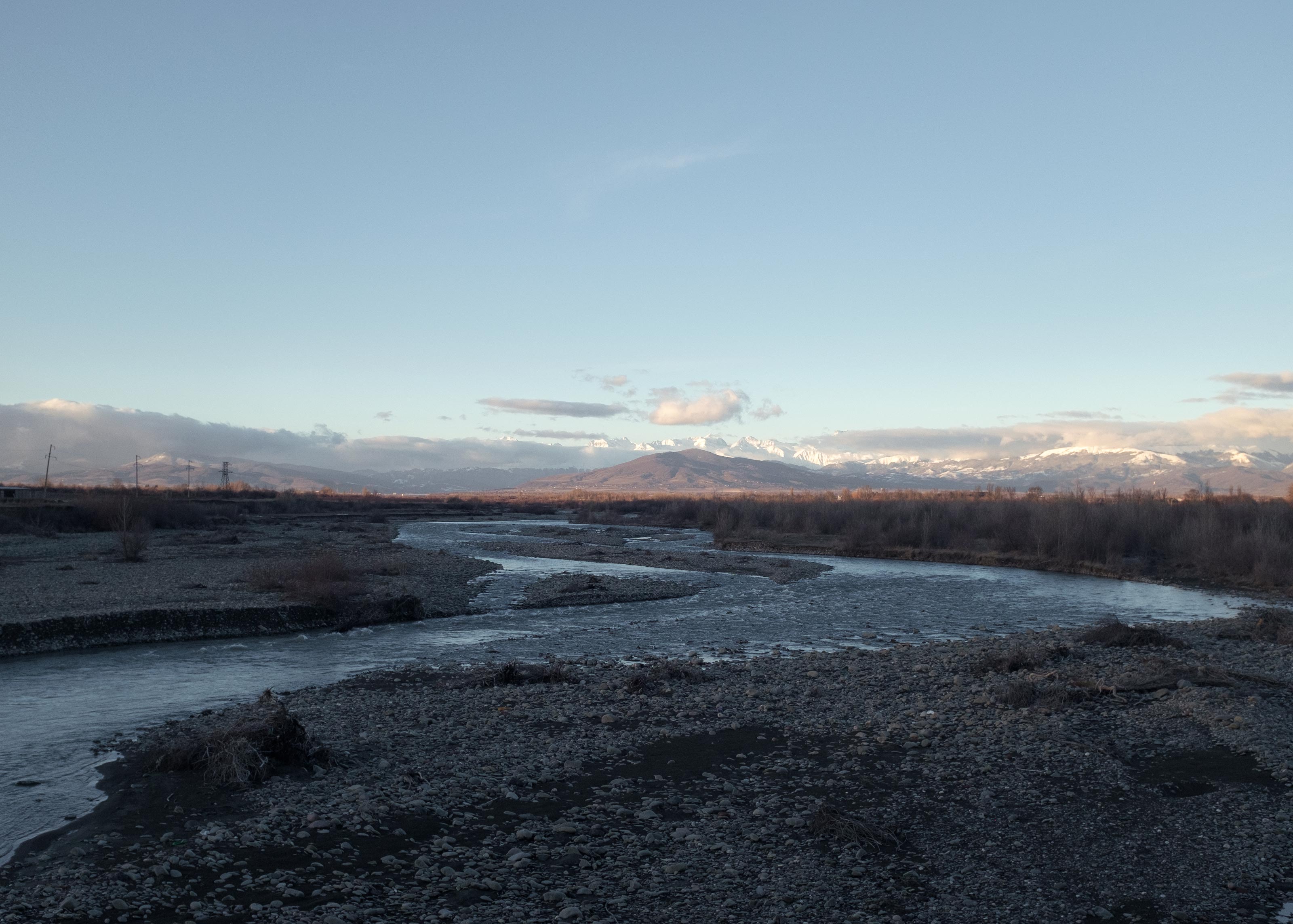 A winding river flows through a rocky, leafless plain under a clear blue sky. In the distance, snow-covered mountains rise behind a line of bare trees in the region.