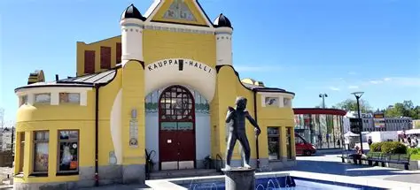 A man wearing glasses and a blazer working at a desk, writing in a notebook beside a computer monitor in a modern office.