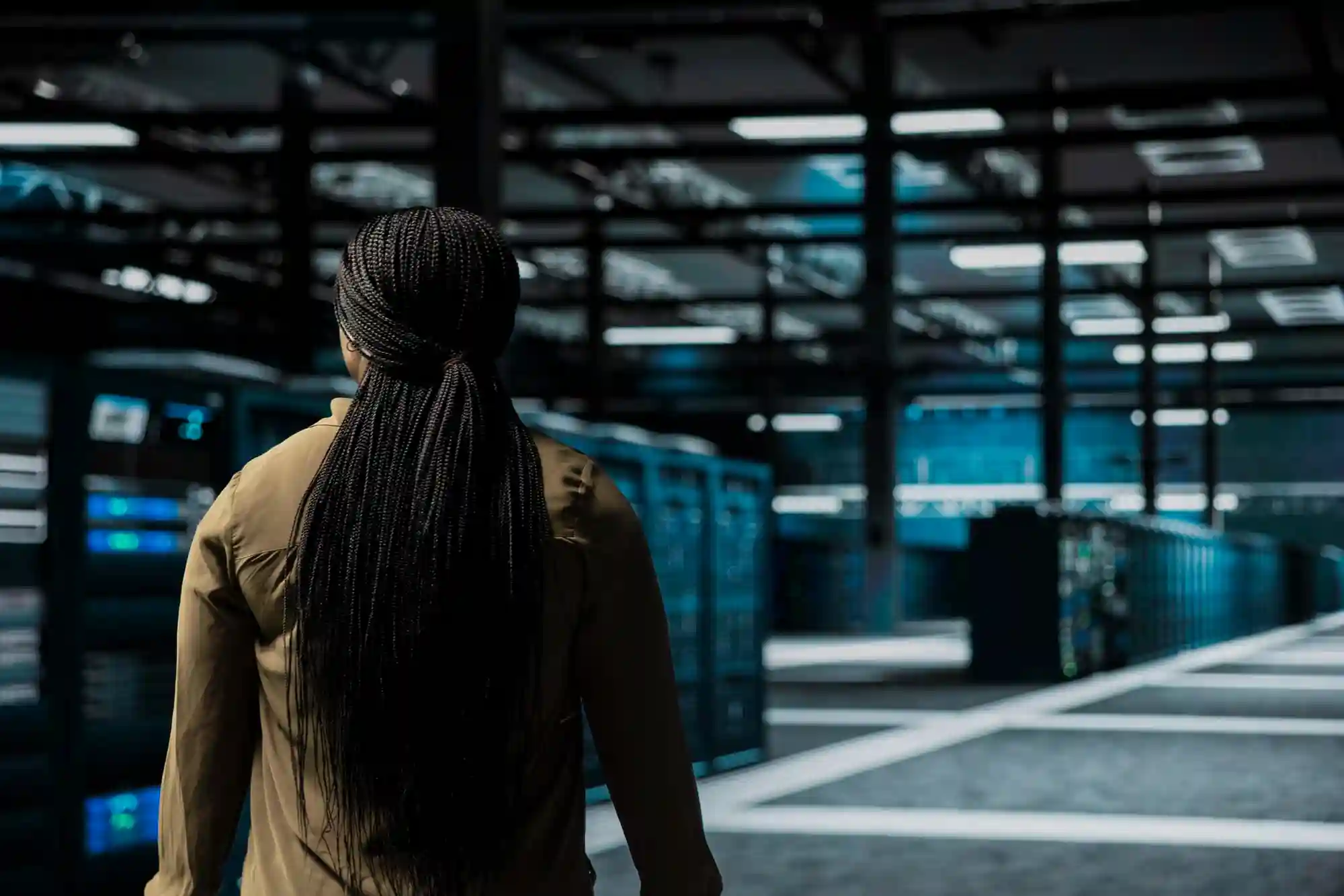 A woman with long dark braids stands in a dimly lit, high-tech server room looking toward rows of equipment.