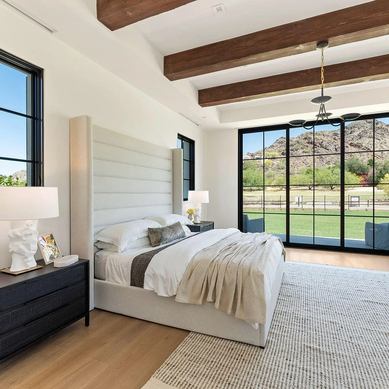 Serene bedroom with a white bed, beige bedding, wooden beams, and large glass doors opening to a scenic outdoor view.