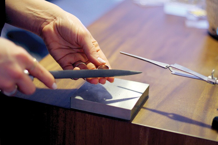 A pair of hands holds a knife and a small metal object over a rectangular metal block on a dark wooden surface. A pair of metal tweezers lies nearby on the block. The background is out of focus.