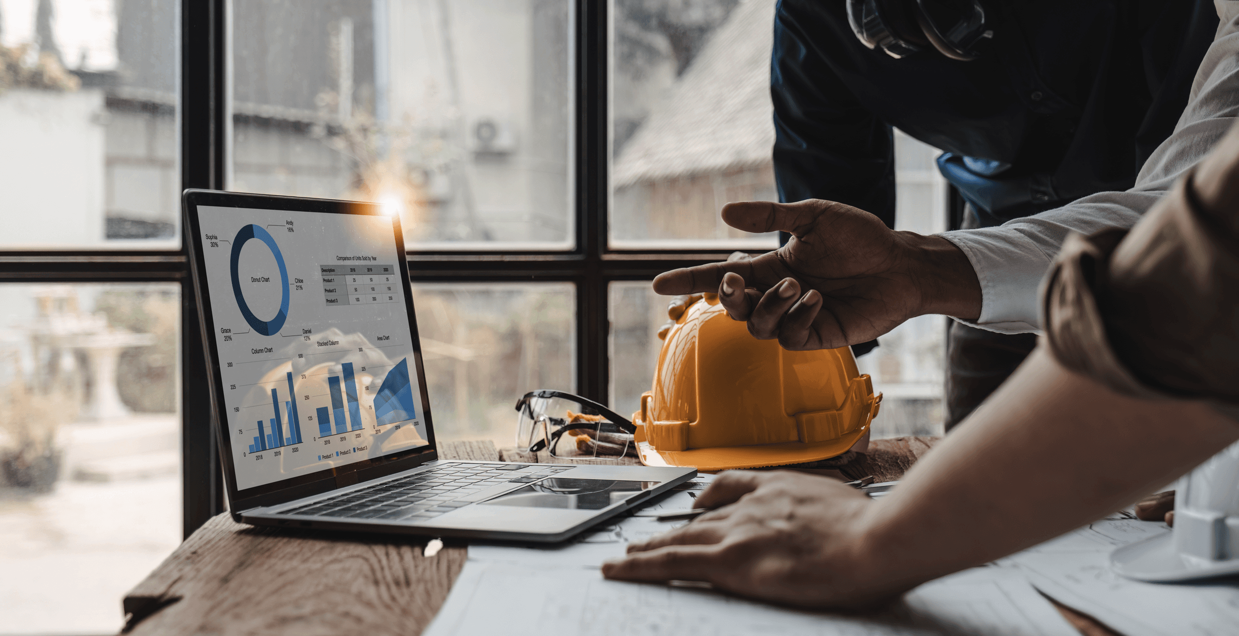 Ingénieurs de TCC Especialização discutant des graphiques de performance sur un ordinateur portable lors d'une réunion de planification de projet de construction. Un casque de sécurité jaune en évidence sur la table, représentant l'accent mis sur la sécurité et l'efficacité dans la gestion des projets.