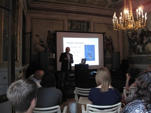 A photo of a man giving a lecture while sitting in a renaissance era room with a screen projector behind him.