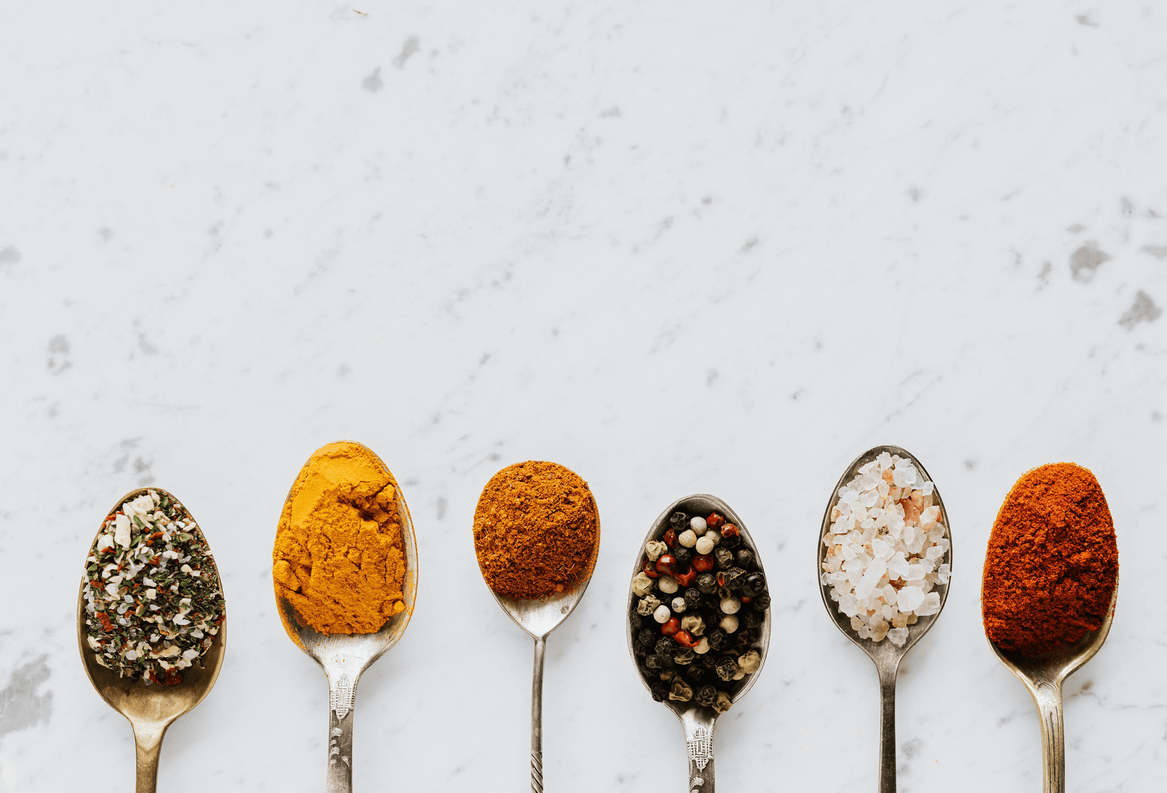 Six vintage spoons lined up on a white marble surface, each filled with a different seasoning — mixed herbs, turmeric, curry powder, mixed peppercorns, pink Himalayan salt, and paprika.