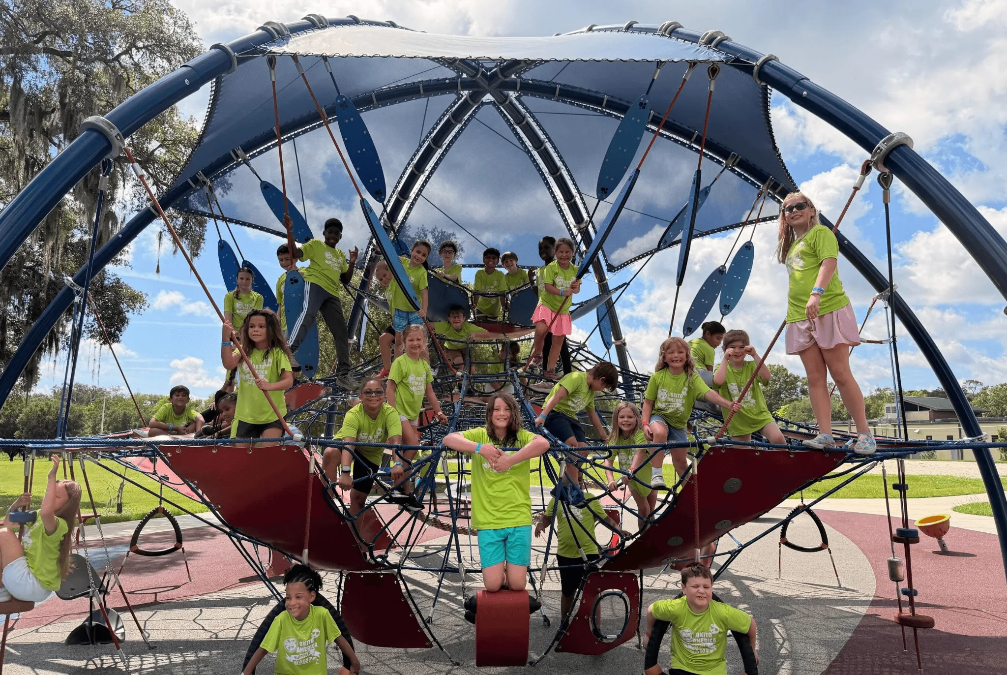 Large group of children wearing bright green shirts posing together on a towering blue and red playground structure with arched canopy and climbing elements under a partly cloudy sky.