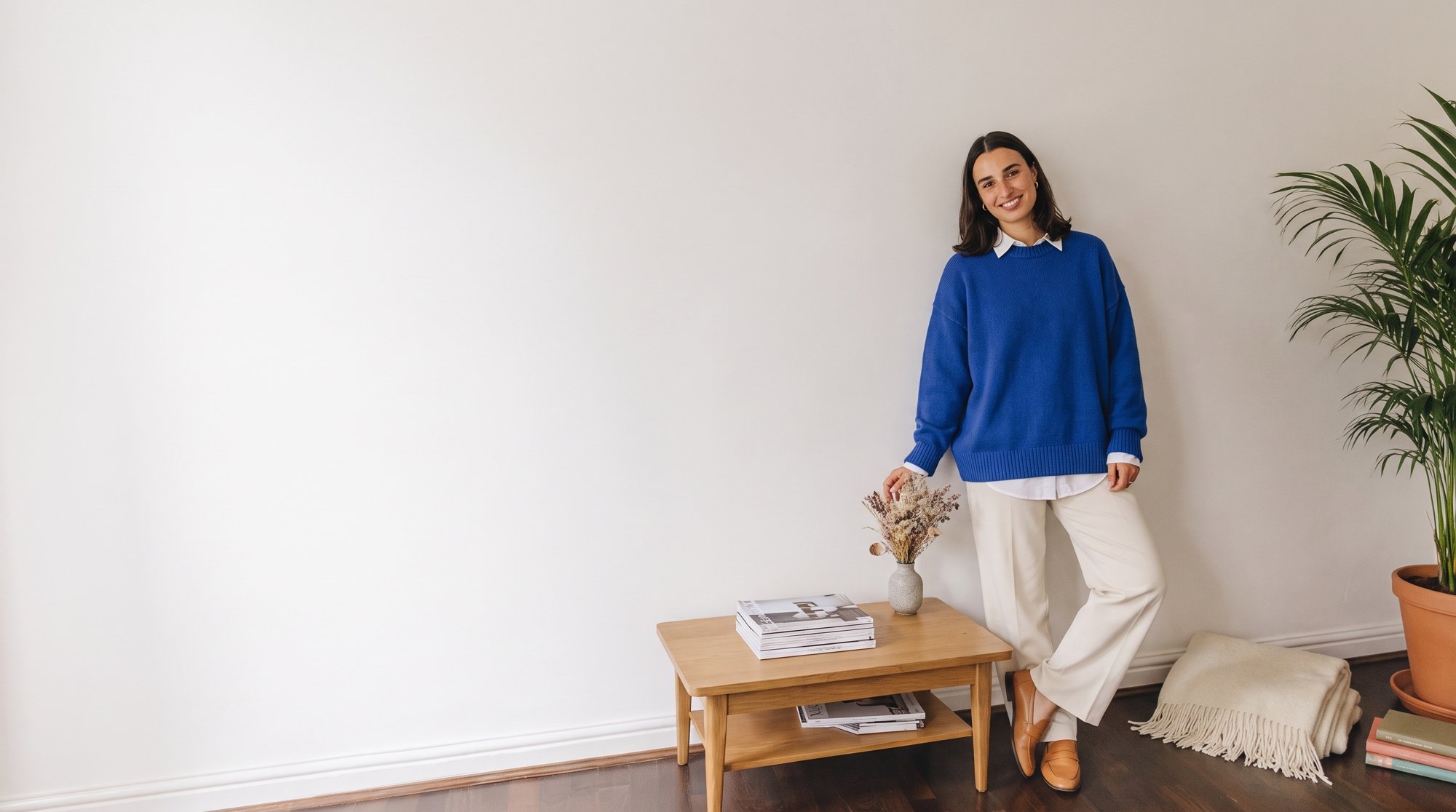 Grace Ellery standing beside a small oak side table in her home office, wearing a blue knit jumper and cream trousers.