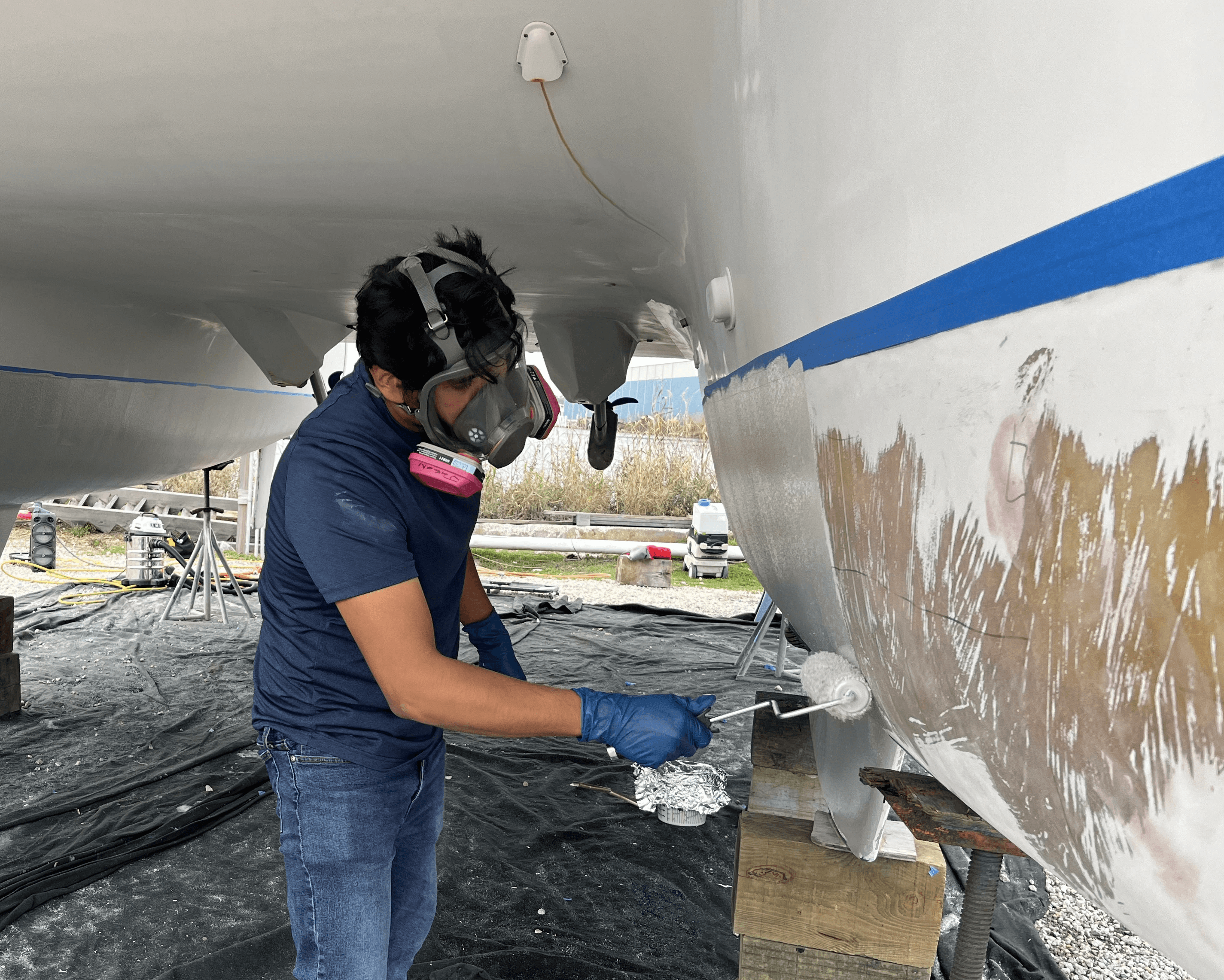 Man in a resperator with gloves on. He is painting the bottom of a boat light grey with a rolling blush. 