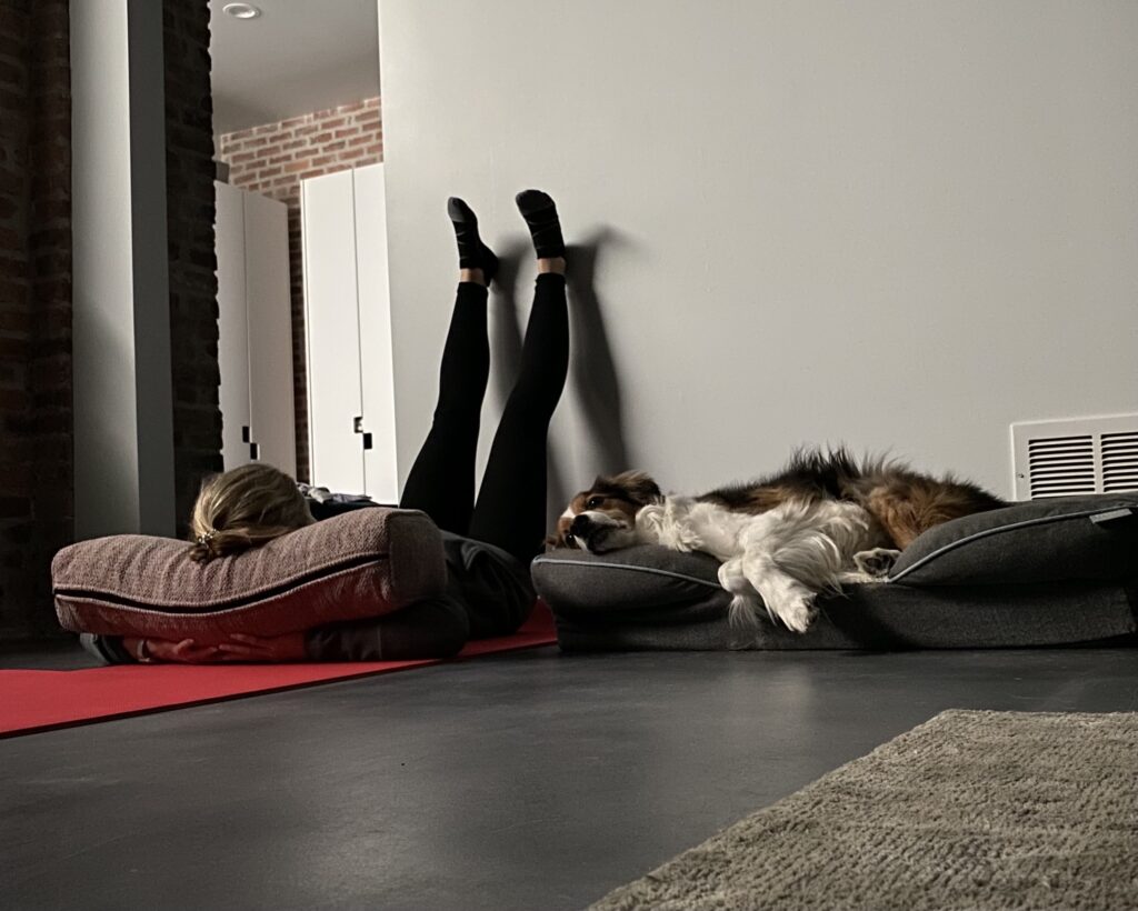 Heather Hausman lies on a yoga mat with legs propped against a wall, while a sleeping dog rests on a nearby cushion in a modern living room featuring a brick wall.