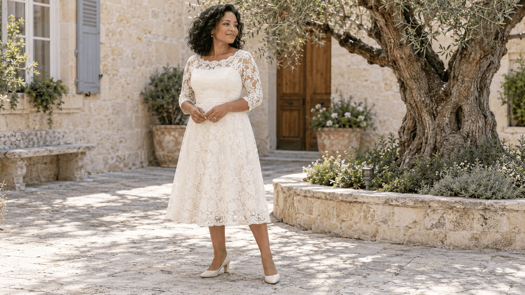 Une femme porte une robe midi ivoire en dentelle doublée, avec des manches trois-quarts, dans une cour arborée.