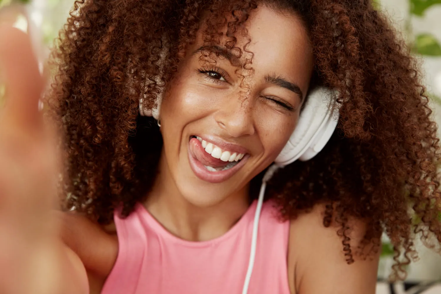 A young person with curly hair smiles and waves at the camera, wearing headphones and a pink shirt.