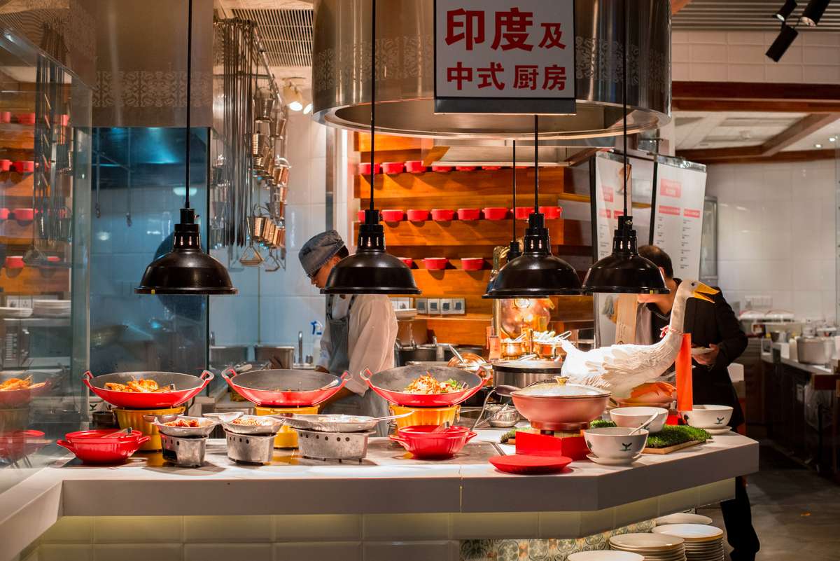 Cooks in white uniforms prepare food in large woks and pots over a long counter under hanging metal lamps and signs.