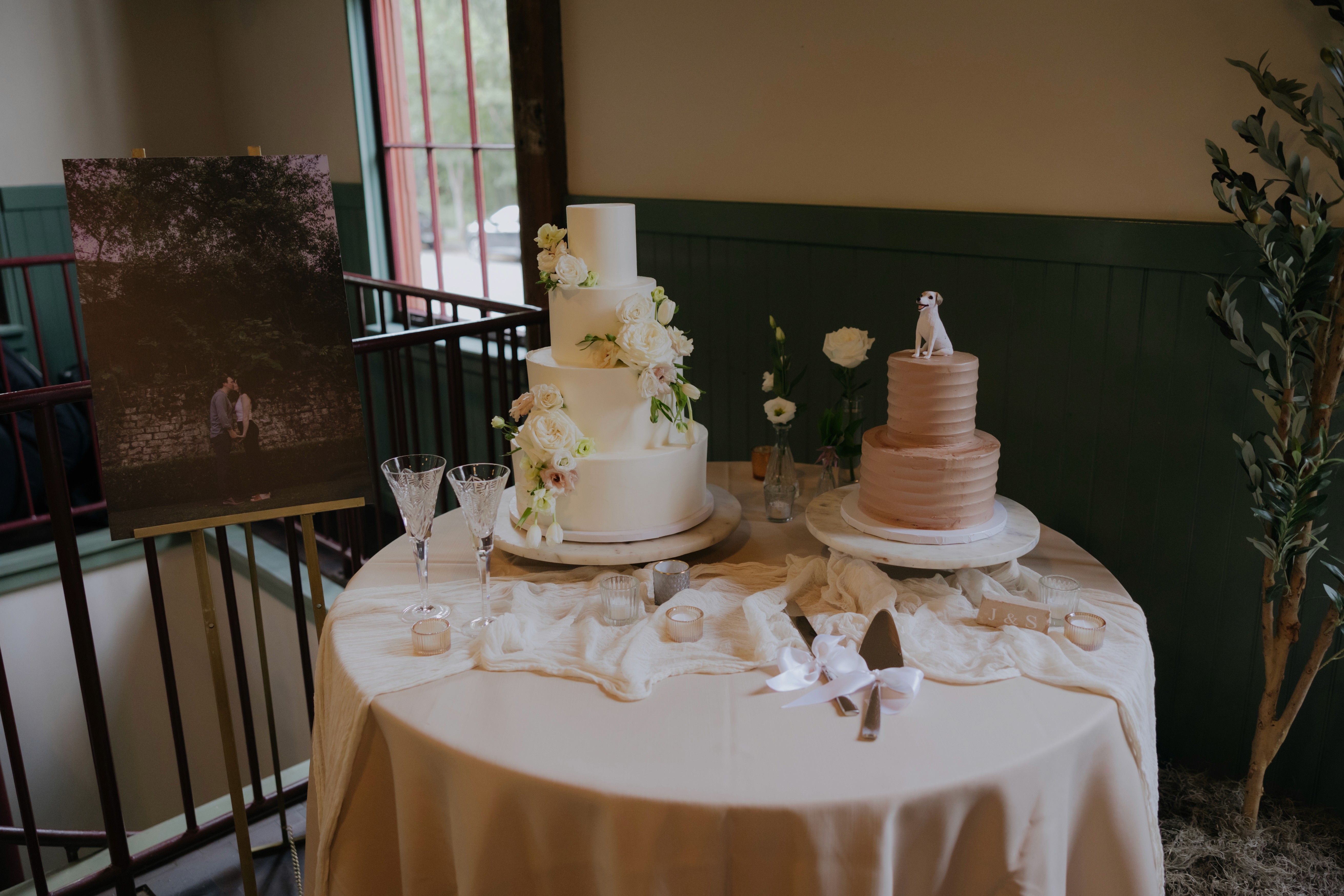 White floral wedding cake displayed beside framed artwork and greenery indoors.